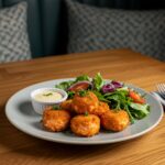 Final presentation of crispy Bang Bang salmon bites on a rustic wooden table alongside a garden salad and dipping sauce, showcasing a delightful meal.