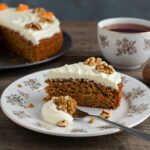 A delicious slice of easy oatmeal carrot cake showcasing its moist texture and cream cheese frosting, elegantly presented on a wooden table next to a steaming cup of tea.