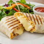Delicious grilled cheese burrito cut in half on a plate, showcasing melted cheese, accompanied by fresh salad and dipping sauce.
