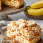 A slice of Banana Pudding Crunch Cheesecake on a white plate, showcasing a creamy, layered dessert with a golden graham cracker crust, topped with whipped cream, slices of ripe banana, and a crunchy crumble topping. The cheesecake has a smooth texture with visible banana slices and a glossy finish, beautifully presented with a sprinkle of crushed cookies around the base.