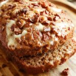A golden-brown loaf of moist banana bread sliced to reveal a tender, dense crumb with specks of mashed bananas. The bread rests on a rustic wooden cutting board, with a few slices fanned out to showcase its texture. Soft crumbs are visible, with a slightly glossy crust that indicates moisture. The background is softly blurred, highlighting the inviting, homemade appeal.