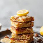 A close-up of vibrant lemon brownies arranged neatly on a white plate, showcasing their golden-brown top glazed with a glossy lemon glaze and flecks of zest. The brownies appear moist, dense, and fudgy with a bright yellow citrus drizzle adding contrast.