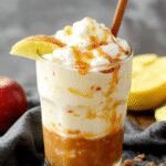 A tall glass filled with a caramel-colored apple cider float topped with whipped cream and drizzled with rich caramel sauce. The drink contains visible apple slices and a cinnamon stick garnish, with a caramel swirl pattern on the surface, set against a rustic wooden table.