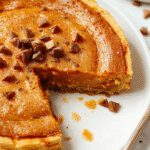 A slice of crustless pumpkin pie on a white plate, topped with a dollop of whipped cream and a sprinkle of cinnamon, set against a warm wooden table