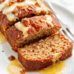 A freshly baked banana cake with a golden-brown crust, sliced to reveal moist, tender layers dotted with ripe banana pieces and a hint of caramelized sugar topping, elegantly plated on a white cake stand with a few banana slices and a sprinkle of powdered sugar for garnish.