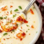 A steaming bowl of white lasagna soup garnished with freshly chopped parsley and shredded mozzarella cheese, showing creamy Alfredo sauce, tender lasagna noodles, and bits of cooked chicken in a white ceramic bowl on a rustic wooden table.