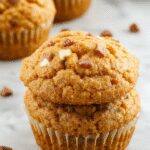 A close-up of golden-brown pumpkin muffins topped with a sprinkle of cinnamon and a dollop of cottage cheese, arranged on a rustic wooden plate with a few pumpkin seeds scattered around. The muffins have a slightly cracked surface revealing moist, fluffy interiors with visible specks of pumpkin and cottage cheese, complemented by a cozy, warm-toned background.