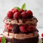 A slice of dark chocolate raspberry mousse cake on a white plate, showcasing a glossy dark chocolate top layer adorned with fresh raspberries and a dusting of cocoa powder, with a rich, creamy interior visible from the cut edge. The background features a blurred elegant table setting with additional fresh raspberries and a drizzle of chocolate sauce.
