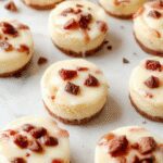 A close-up of several mini cheesecakes arranged on a marble platter. Each cheesecake has a golden graham cracker crust, creamy pale yellow filling, and is topped with a swirl of whipped cream and a fresh strawberry. The mini desserts are uniformly sized, with smooth textures and a glossy finish, invitingly presented for a casual yet elegant dessert display.