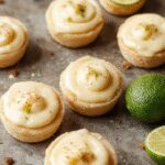 A close-up view of several mini key lime pies arranged on a elegant white platter. The pies have golden graham cracker crusts filled with vibrant lime filling, topped with a dollop of whipped cream and garnished with a tiny lime wedge. The background is softly blurred, emphasizing the glossy, smooth texture of the lime filling and the crisp crusts.