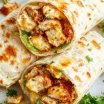 A close-up of a cheesy garlic chicken wrap sliced in half, revealing melted cheese, golden grilled chicken pieces, fresh lettuce, and tomato slices inside a soft flour tortilla. The wrap is presented on a rustic wooden plate with a slight steam, highlighting its gooey texture and vibrant ingredients.