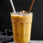 A tall glass filled with a creamy, amber-colored pumpkin chai iced latte, topped with a dollop of whipped cream and a sprinkle of cinnamon. The drink showcases a layered appearance with visible swirls of pumpkin and chai flavors, garnished with a cinnamon stick and a few pumpkin seeds on the side. The background features a rustic wooden table with autumnal decor, enhancing the cozy, inviting aesthetic.
