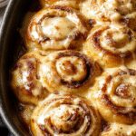 A golden-brown pumpkin cinnamon roll bake in a rectangular ceramic dish, topped with a drizzle of creamy glaze and sprinkled with cinnamon. The bake appears moist and fluffy, with visible swirls of pumpkin and cinnamon throughout.