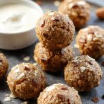 Close-up of golden-brown pumpkin protein balls arranged in a neat cluster on a rustic wooden plate, garnished with a sprinkle of cinnamon and tiny pumpkin seeds, with a blurred fall-themed background.