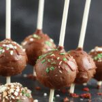 A close-up of a Starbucks-style cake pop coated in smooth white icing with colorful sprinkles on top, presented on a small plate with a blurred coffee cup in the background, highlighting the glossy texture and festive decoration.