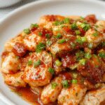 A close-up of tender, golden-brown chicken pieces coated in a glossy, vibrant red sweet chili glaze garnished with sliced green onions and sesame seeds on a white plate, with a background of fresh ingredients and a dipping bowl of extra sauce.