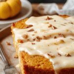 A slice of pumpkin bread topped with a swirl of vanilla pudding, served on a rustic wooden plate. The bread has a golden-brown crust with visible pumpkin specks, complemented by a creamy white vanilla pudding glaze that drips slightly over the edges. The presentation is enhanced with a light dusting of powdered sugar and a sprig of fresh mint for contrast.