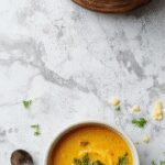 A vibrant bowl of vegan lemon lentil soup with a bright yellow hue, garnished with fresh herbs and a lemon wedge, served on a rustic wooden table with a spoon nearby, steam gently rising from the soup.