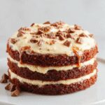 A layered white German chocolate cake with smooth white frosting, topped with toasted coconut flakes and pecans, presented on a white platter with a decorative fork. The cake slices reveal a moist, fluffy interior contrasted by the glossy icing.