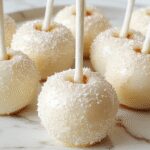 A close-up of elegant white candy apples coated with a glossy, smooth white glaze, each topped with a delicate edible glitter sparkle. The apples are presented on a decorative tray with a soft-focus background featuring subtle winter decor and twinkling lights, emphasizing their pristine appearance and festive charm.