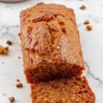 A slice of golden-brown pumpkin spice bread with a slightly cracked top, topped with a sprinkle of cinnamon and a drizzle of glaze. The bread is presented on a rustic white plate with a few pumpkin seeds and a cinnamon stick beside it, set against a warm wooden table background.