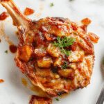 Plate featuring two golden-brown pork chops topping a glossy, ruby-red apple chutney with visible chunks of apple and onion. The meat rests on a rustic wooden surface, with a garnish of fresh herbs and a side of caramelized apples adding visual appeal.