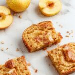 A golden-brown loaf of apple cinnamon bread on a rustic wooden cutting board, sliced to reveal a moist interior with visible chunks of apples and cinnamon swirls. The bread has a glossy sugar glaze on top, with a few slices fanned out beside it. Soft natural light highlights the textures and warm tones of the bread and apples.