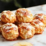 Golden-brown apple fritter bites served on a white plate, dusted with powdered sugar, with visible chunks of apple and a crispy textured surface, garnished with a sprig of mint.