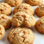 Golden-brown apple snickerdoodles arranged on a rustic white plate with cinnamon sugar coating visible. The cookies have a slightly cracked surface, revealing a soft interior, garnished with a sprinkle of cinnamon. The background includes fall-themed elements like a wooden table, autumn leaves, and a cinnamon stick, creating a warm, inviting atmosphere.