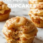 A close-up of golden brown apple pie cupcakes in individual paper liners, topped with a sprinkle of cinnamon and sugar, showcasing a crispy, flaky crust filled with diced apple filling. The cupcakes are arranged on a rustic wooden surface, with a few slices of fresh apple and cinnamon sticks in the background.