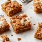 A close-up of frosted carrot cake bars arranged on a rustic wooden platter. The bars are golden orange with a creamy white frosting drizzled on top, speckled with tiny carrot shreds and sprinkled with chopped nuts. The background features a softly blurred kitchen setting with additional cake bars and fresh carrots.