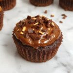 A close-up of two decorated Chocolate Pumpkin Cupcakes with orange and black themed Halloween toppers, glazed with shiny chocolate frosting and garnished with pumpkin-shaped candies, set on a rustic wooden surface with autumn leaves in the background.