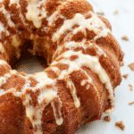 A golden-brown cinnamon swirl Bundt cake with a glossy glaze drizzled over the top, showcasing a beautifully spiral pattern. The cake is displayed on a elegant white plate with a few cinnamon sticks and powdered sugar sprinkled around. The textured surface of the cake highlights its moist crumb and cinnamon streaks, inviting a cozy baking vibe.