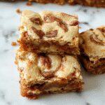 A close-up of a freshly sliced cinnamon swirl snickerdoodle blondie on a rustic wooden plate, showcasing its golden-brown crust with a cinnamon caramel swirl pattern, topped with a light dusting of powdered sugar. The background features a cozy kitchen setting with warm lighting and a few cinnamon sticks and baking utensils artfully arranged.