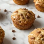 A close-up of golden-brown coffee cake muffins arranged on a rustic wooden tray. The muffins have a crumbly streusel topping with a hint of cinnamon, and they are presented with a few fresh coffee beans scattered around. The muffins are in paper liners, and the texture looks moist and tender with a crispy crumbly topping.