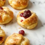 Close-up of golden-brown crescent-shaped bites topped with vibrant red cranberry sauce and creamy white cheese, arranged on a white plate garnished with fresh rosemary, with a hint of fall-themed decor in the background