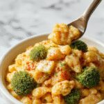 A creamy, golden bowl of broccoli cheddar orzo sits atop a rustic wooden table. The dish is garnished with fresh chopped herbs, with tender broccoli florets and perfectly cooked orzo visible, showcasing a rich, cheesy sauce that glistens. The presentation features a cozy setting with a spoon partially inserted into the bowl, highlighting its velvety texture and hearty appeal.
