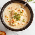 A bowl of creamy chicken parmesan soup featuring chunks of tender chicken, a rich, cheesy sauce with a sprinkle of herbs, garnished with fresh basil leaves. The soup sits in a rustic white bowl on a wooden surface, with a spoon resting nearby, highlighting its creamy texture and hearty ingredients.