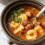 A steaming bowl of cheesy tortellini soup garnished with fresh herbs, served in a rustic white bowl on a wooden surface. The soup contains plump tortellini, diced sausage, and vibrant vegetables, with a golden broth that looks rich and flavorful. The scene is complemented by a background of ingredients and a cozy kitchen setting, emphasizing warmth and homemade comfort.