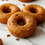 A close-up of golden-brown baked pumpkin donuts arranged on a rustic ceramic plate, topped with a light dusting of powdered sugar and sprinkled with cinnamon. The donuts showcase a tender, moist interior with a slight sheen, highlighting their flavorful pumpkin and spice aroma. The background features autumnal elements like small pumpkins, cinnamon sticks, and a cozy checkered cloth, creating a warm, seasonal fall vibe.