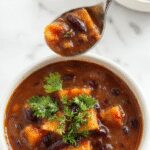 A vibrant bowl of black bean and sweet potato soup garnished with fresh cilantro, served with a spoon on a rustic wooden table. The soup has a rich orange color with black beans visible throughout, and steam rises from the hot dish, suggesting warmth and comfort.
