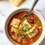 A steaming bowl of Ravioli Soup featuring plump cheese-filled ravioli, vibrant green spinach, slices of Italian sausage, and a rich broth garnished with grated cheese and fresh herbs, set on a rustic wooden table with a soup spoon and bread in the background.
