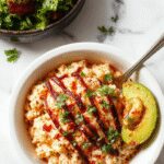A vibrant taco rice bowl featuring fluffy white rice topped with seasoned ground beef, melting cheese, fresh chopped tomatoes, avocado slices, shredded lettuce, and a sprinkle of cilantro. The colorful ingredients are arranged in a rustic bowl with a side of lime wedges, creating an appealing and appetizing presentation.