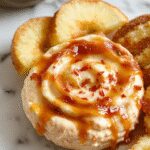 A creamy caramel apple cheesecake dip served in a clear glass bowl, topped with sliced fresh apples and drizzled with caramel sauce. The dip has a smooth, luscious texture with swirls of caramel visible throughout, surrounded by apple slices with a hint of cinnamon on the surface. The background features a cozy fall-themed setting with rustic wooden table and autumn decorations, highlighting a warm and inviting atmosphere.