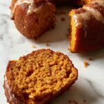 A beautifully baked pumpkin bundt cake with a golden-brown crust, presented on a white cake stand. The cake has a distinct ridged pattern from the 6-cup bundt pan and is topped with a light dusting of powdered sugar. Surrounding the cake are small pumpkin decorations and autumn leaves, creating a festive seasonal display. The background features a rustic wooden table with warm, natural lighting that highlights the cake’s moist texture and inviting appearance.