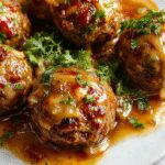 A close-up of golden-brown meatballs glazed with rich onion sauce, served on a white plate. The meatballs are topped with caramelized onions and fresh herbs, creating an appetizing and hearty presentation.