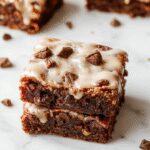 A close-up of a square frosted coffee brownie with a glossy coffee-flavored frosting on top, sprinkled with tiny chocolate chips. The brownie has a rich, fudgy texture with a slightly cracked surface, presented on a rustic wooden plate with a few coffee beans scattered around.