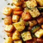 A rustic ceramic platter featuring golden-brown roasted potatoes, vibrant orange carrots, and tender green zucchini slices, all coated with herbs and garlic, arranged attractively with a sprinkle of fresh herbs on top, showcasing crispy edges and fresh ingredients.