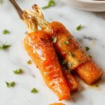 A close-up of a white serving dish filled with glossy, caramel-colored honey glazed carrots. The carrots are sliced diagonally, coated with a shiny honey glaze, and garnished with fresh herbs. The background features a rustic wooden table with hints of holiday décor, evoking warmth and festive cheer.