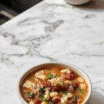 A rustic bowl of ham and bean soup, generously filled with chunks of pink ham, creamy white beans, and vibrant green herbs, set on a wooden table. The soup has a hearty, thick texture with steam gently rising from it, garnished with freshly chopped parsley and a slice of crusty bread on the side.